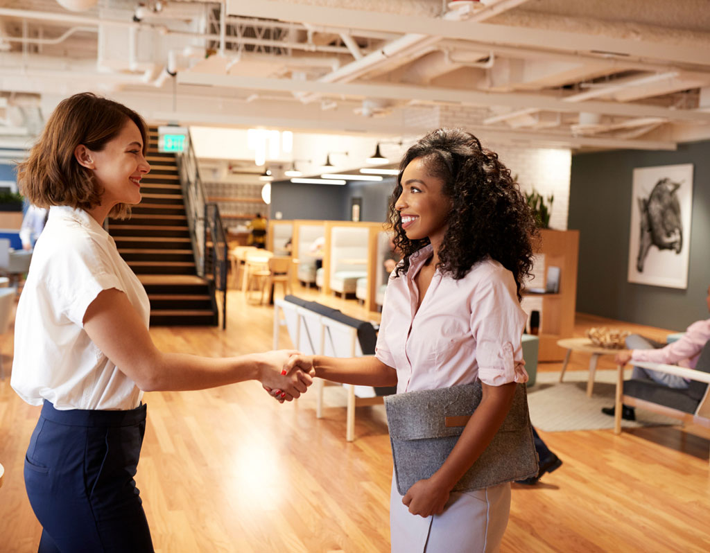 Two Businesswomen Meeting And Shaking Hands In Modern Open Plan Office