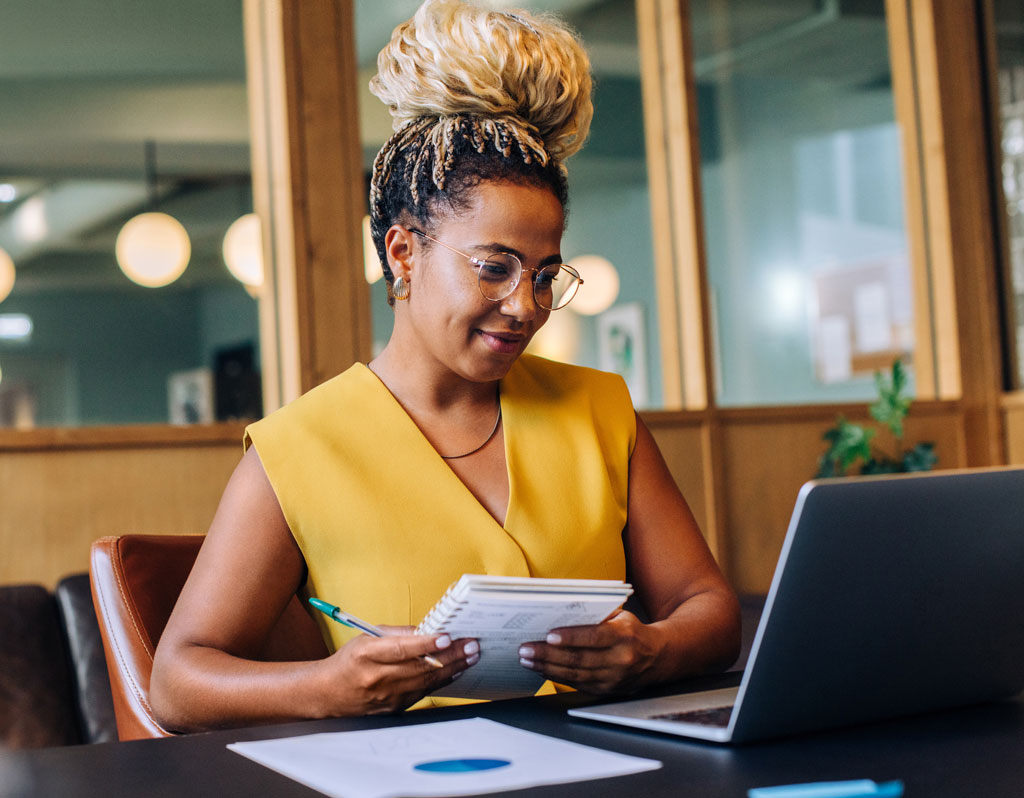 A young black woman, sitting at a desk with a laptop and materials