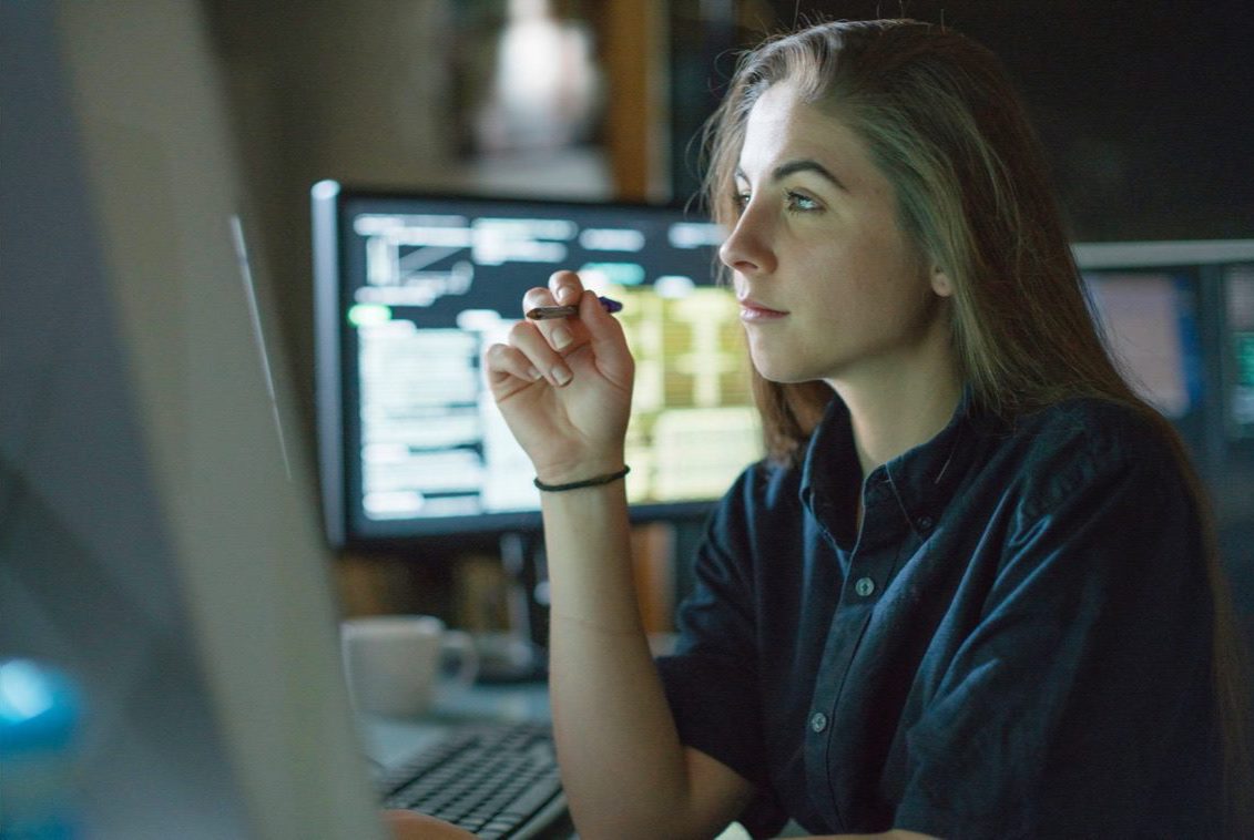 A person sitting at a computer, holding a pen while focusing on a monitor offscreen